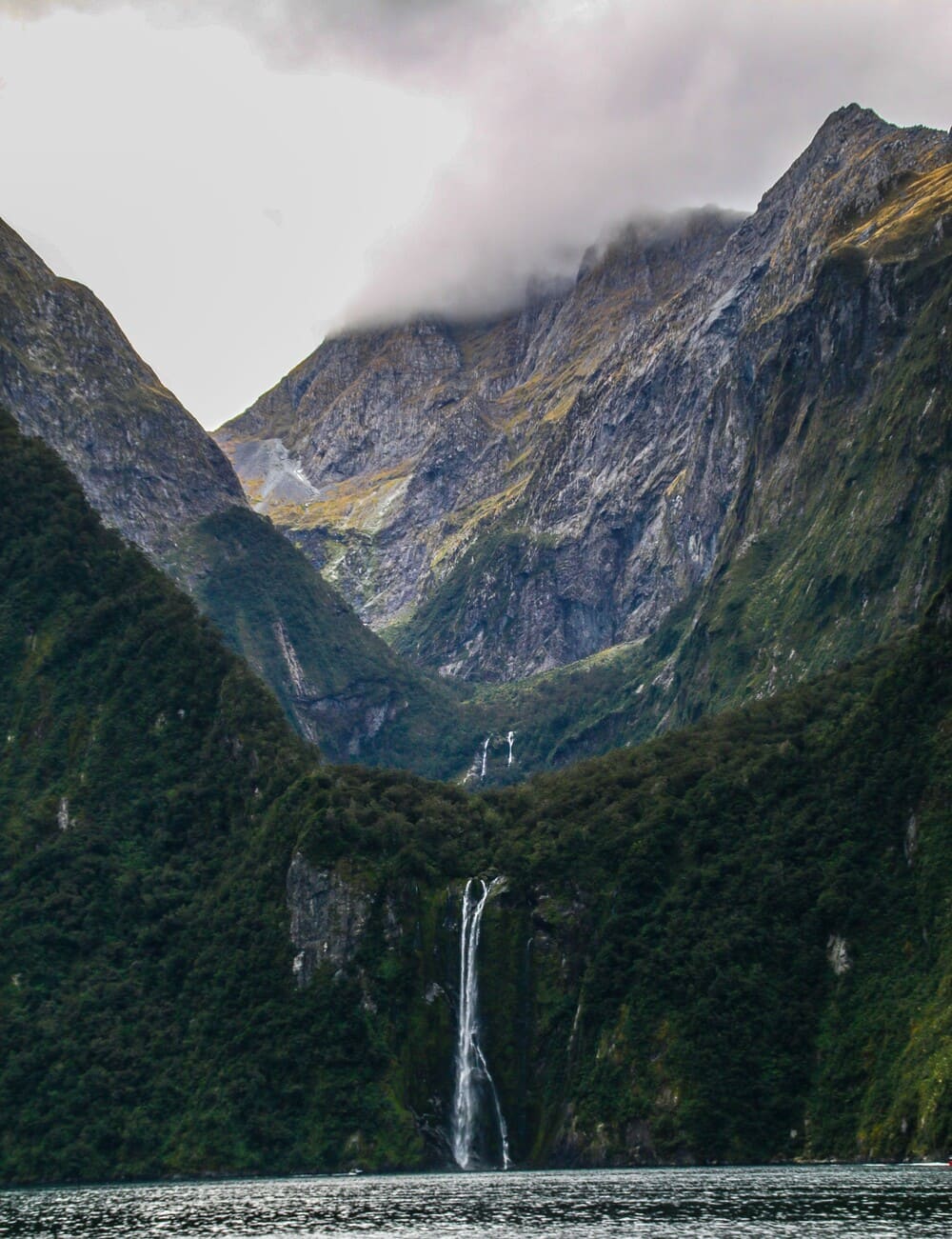 A mountain sticking out of the water, with the bottom covered in plants and a small waterfall on it, in milford sound, New Zealand.